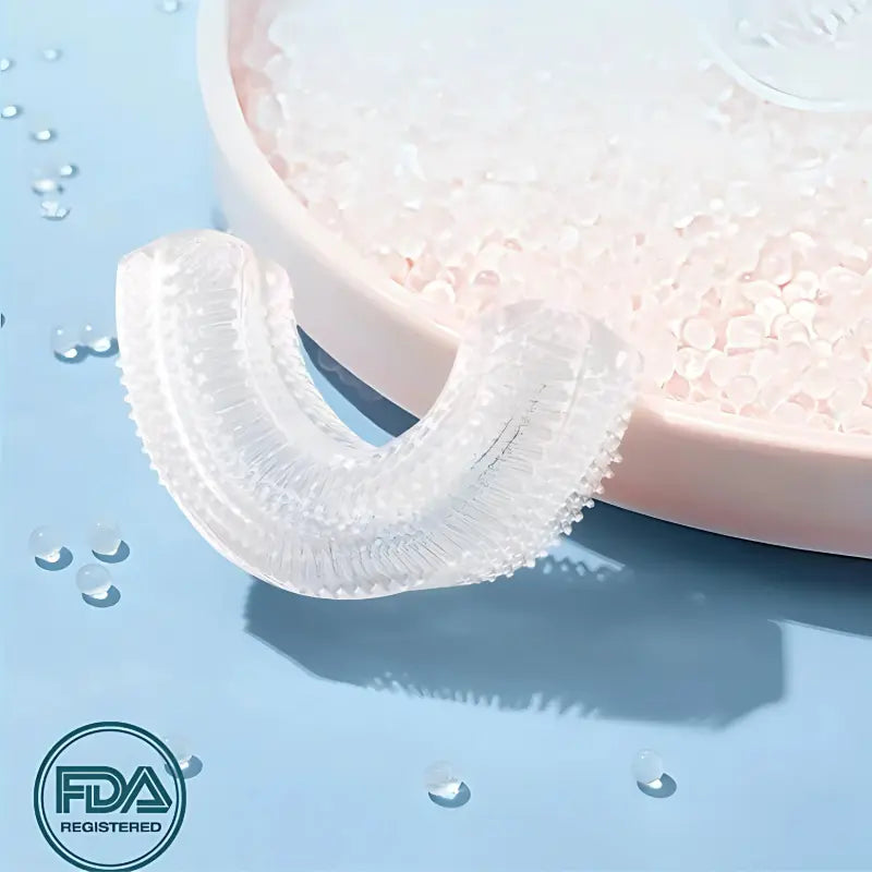Transparent U-shaped replacement toothbrush head with FDA registered silicone material displayed on a blue surface next to small white cleaning beads.